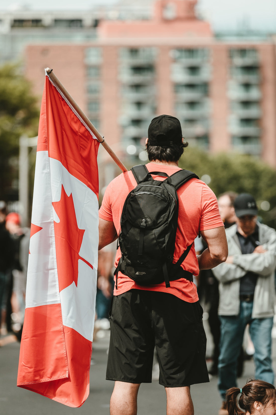 Man in casual clothes with backpack holding Canadian flag outdoors, city background, The Snarky Pirate