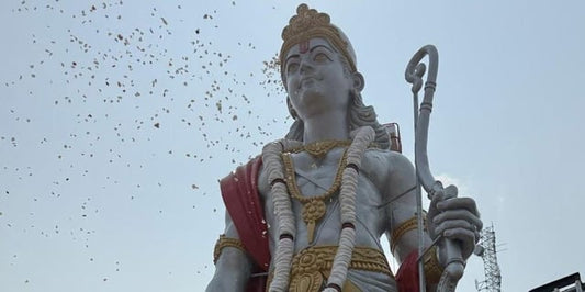 Tall Lord Ram statue outdoors with garlands, red sash, blue sky, and birds flying above